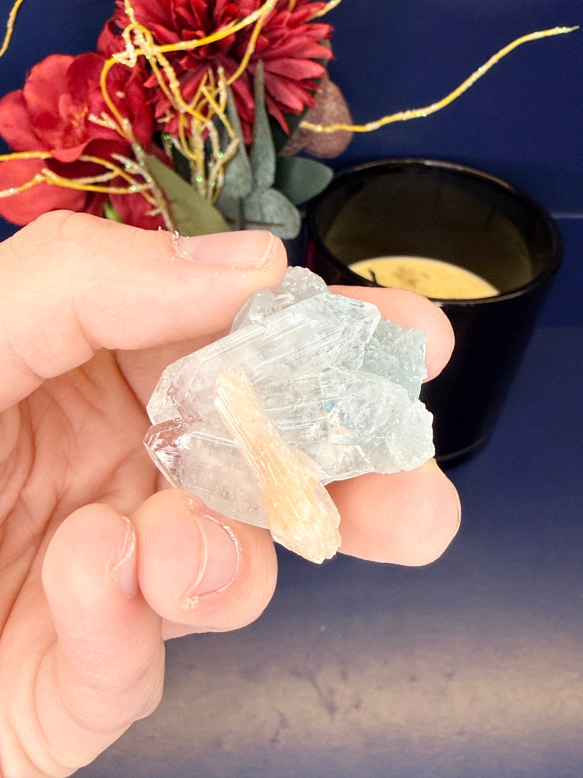 Hand holding a raw green apophylite with peach stillbite crystal in front of a red flower and black candle jar