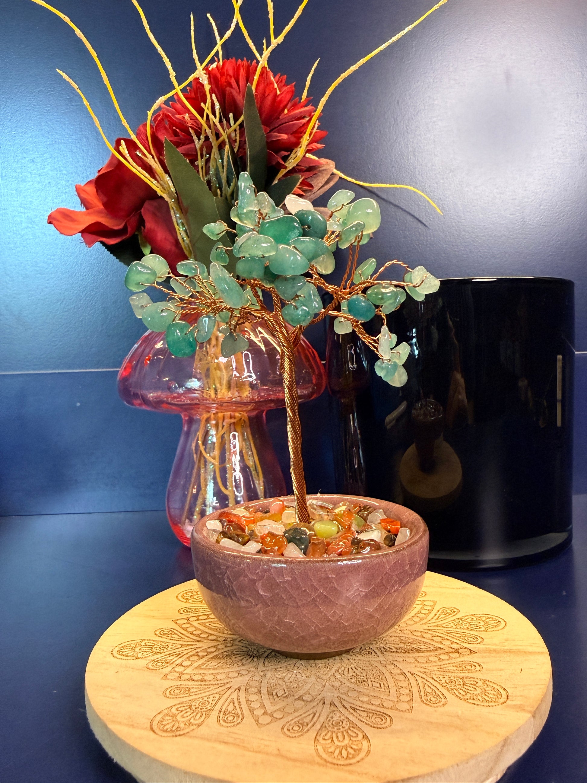 A green crystal chip tree made of copper wire in a pink bowl on a wooden coaster in front of a pink mushroom and black jar