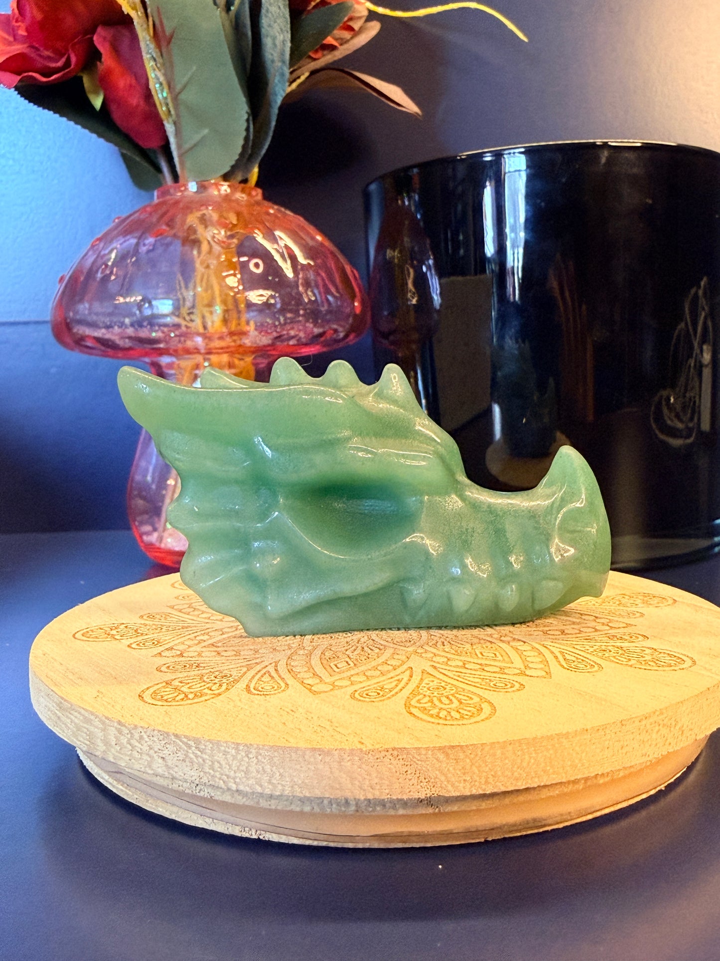 A large green aventurine dragon head sitting on a decorative wooden coaster in front of a glass mushroom and black jar