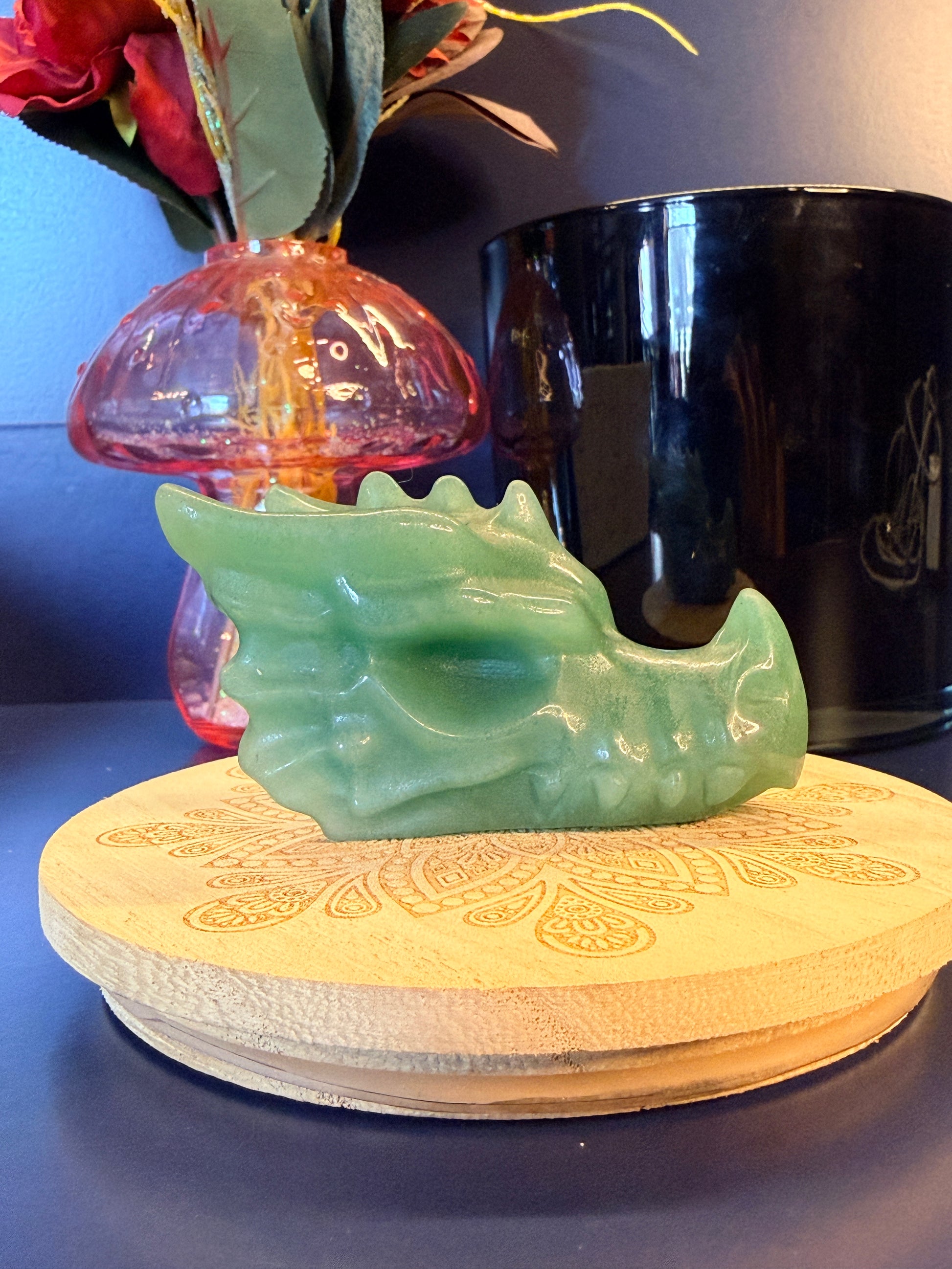 A large green aventurine dragon head sitting on a decorative wooden coaster in front of a glass mushroom and black jar