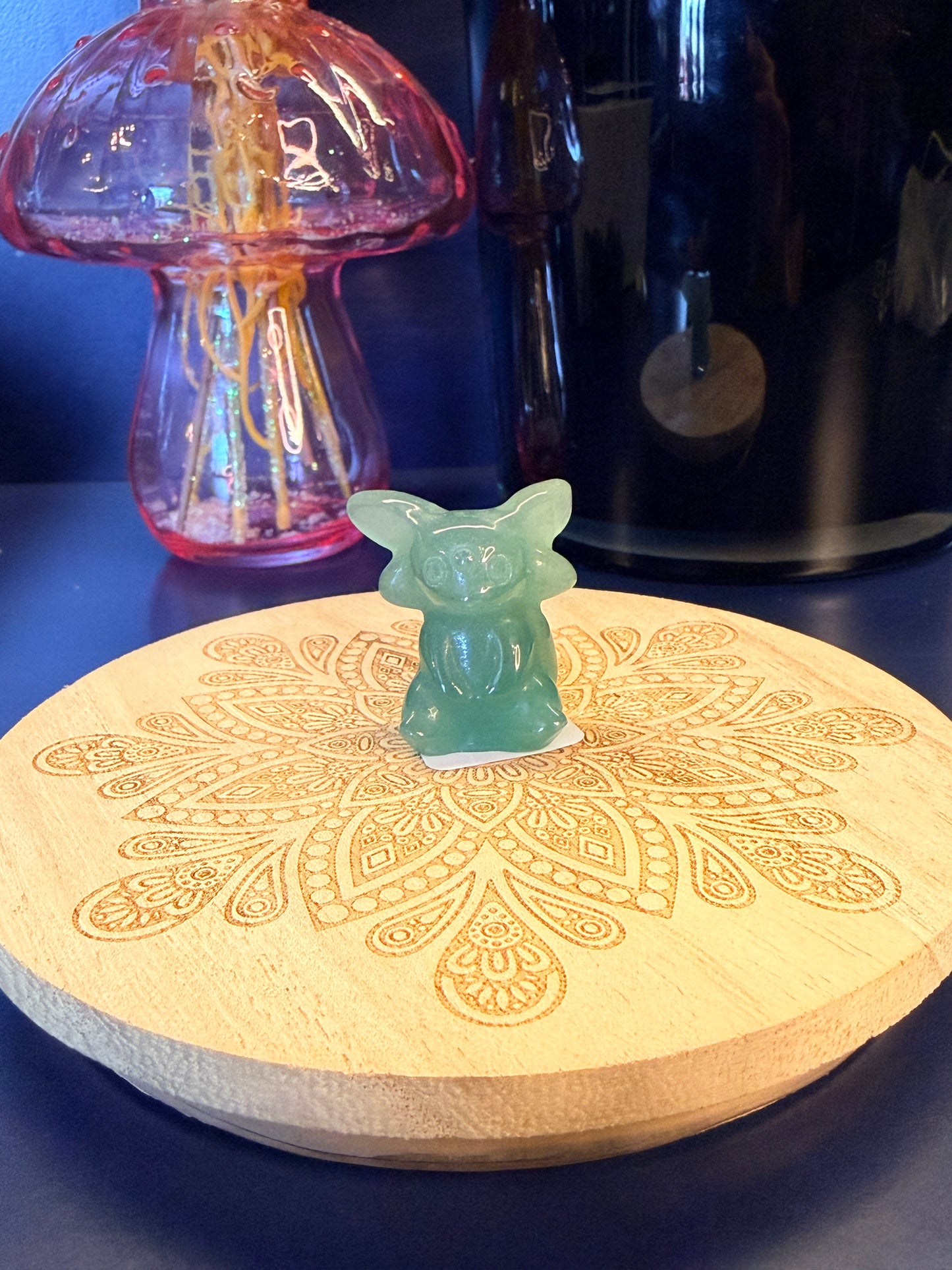 A green crystal espeon sitting on a decorative wooden coaster in front of a glass mushroom and black jar