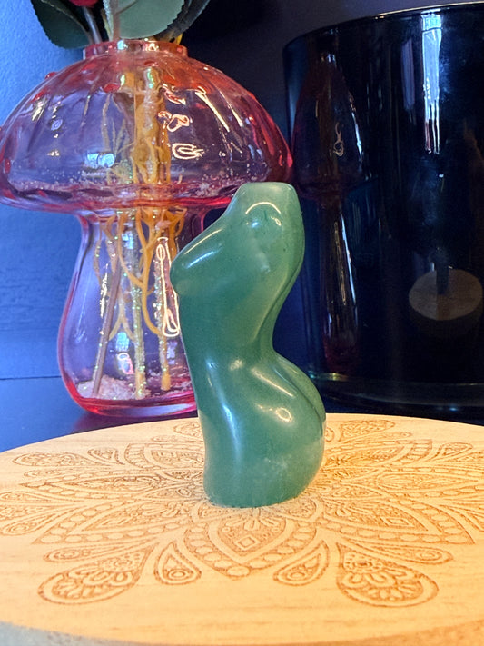 A green crystal goddess body sitting on a decorative wooden coaster in front of a mushroom and black jar