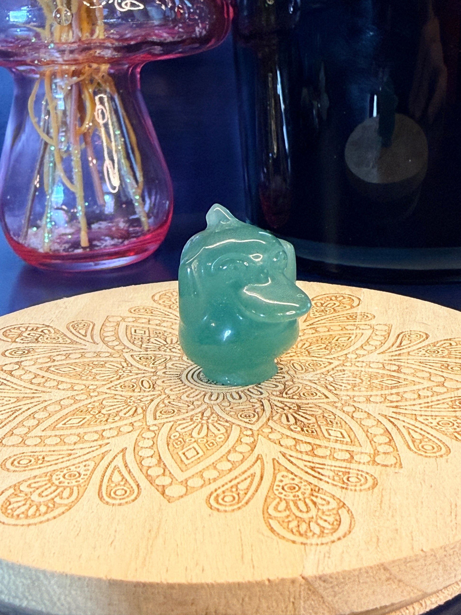 Green Aventurine psyduck-inspired creature sitting on a wooden coaster in front of a black jar and glass mushroom 