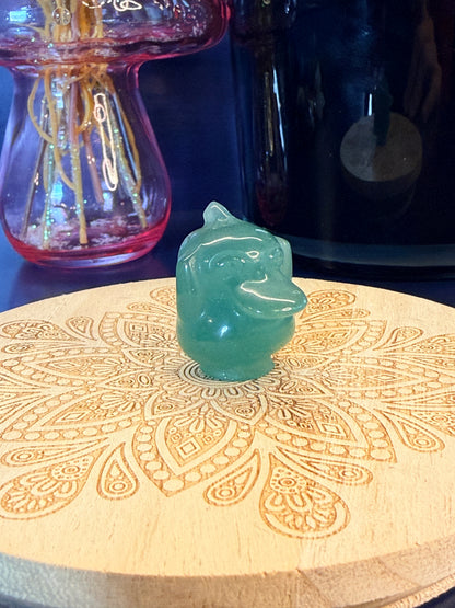 Green Aventurine psyduck-inspired creature sitting on a wooden coaster in front of a black jar and glass mushroom 