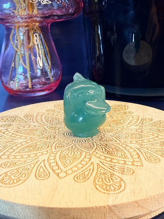 Green Aventurine psyduck-inspired creature sitting on a wooden coaster in front of a black jar and glass mushroom 