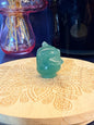 Green Aventurine psyduck-inspired creature sitting on a wooden coaster in front of a black jar and glass mushroom 