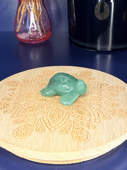 green aventurine turtle carving in front of a pink vase and black jar on a wooden coaster against a blue background