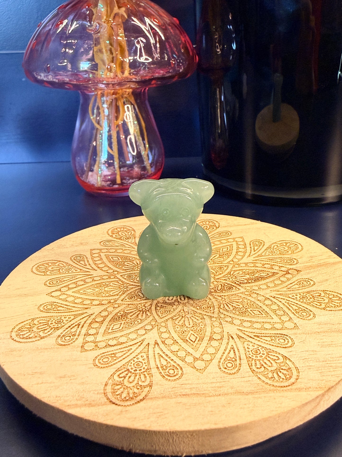 A green aventurine tigger crystal sitting on a decorative wooden coaster in front of a glass mushroom and black jar