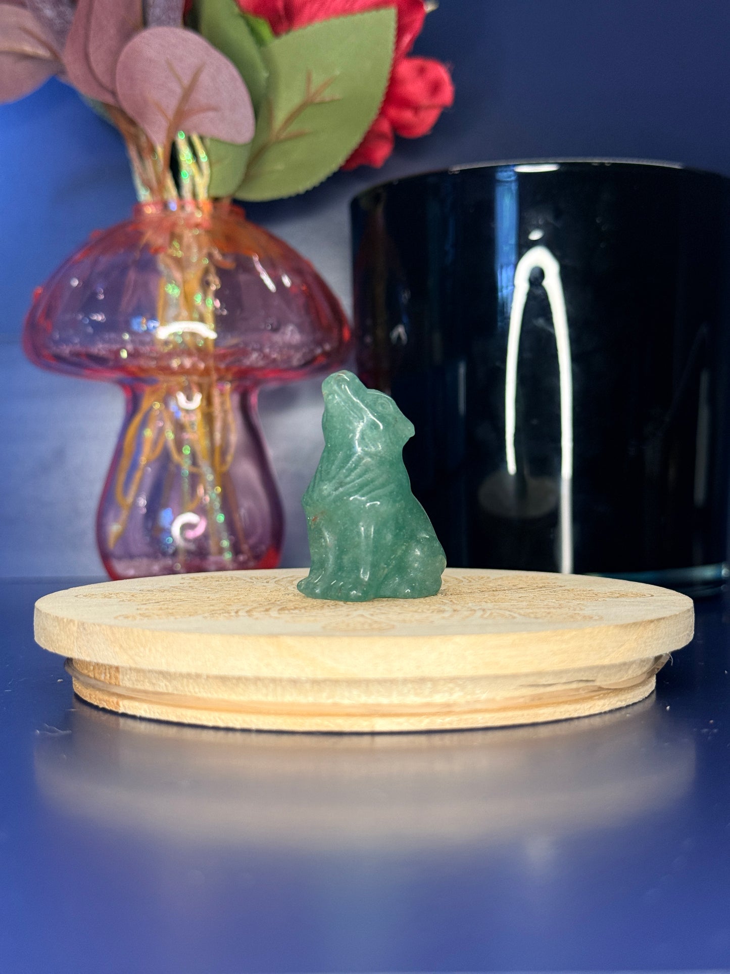 Green Aventurine wolf carving in front of a pink vase and black jar on a wooden coaster against a blue background