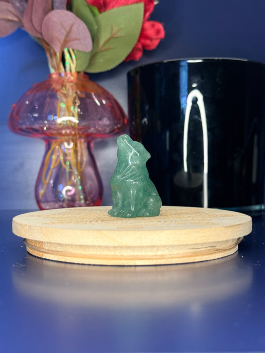 Green Aventurine wolf carving in front of a pink vase and black jar on a wooden coaster against a blue background