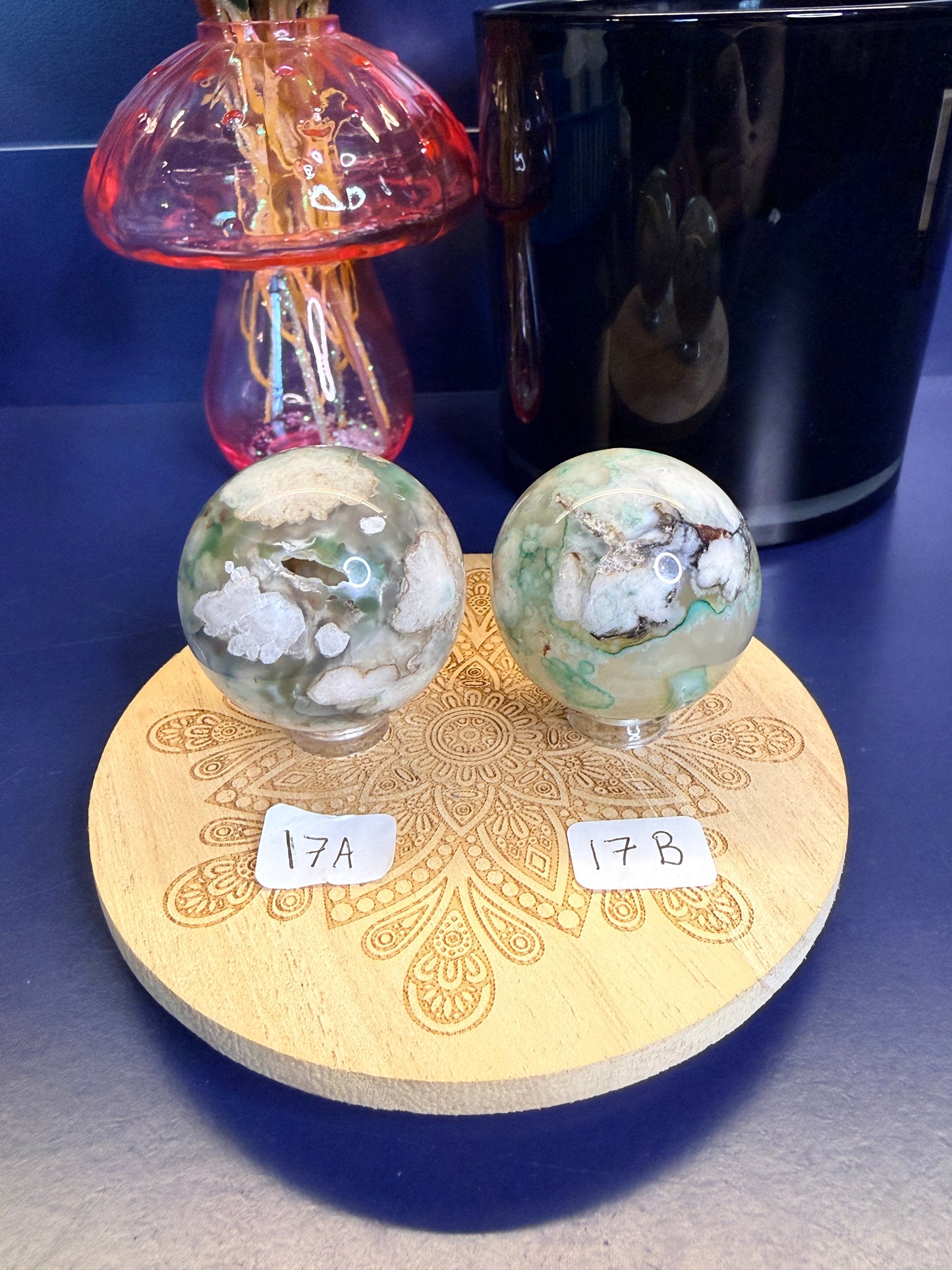 Two white and green translucent flower agate spheres on a wooden coaster in front of a black jar and pink mushroom