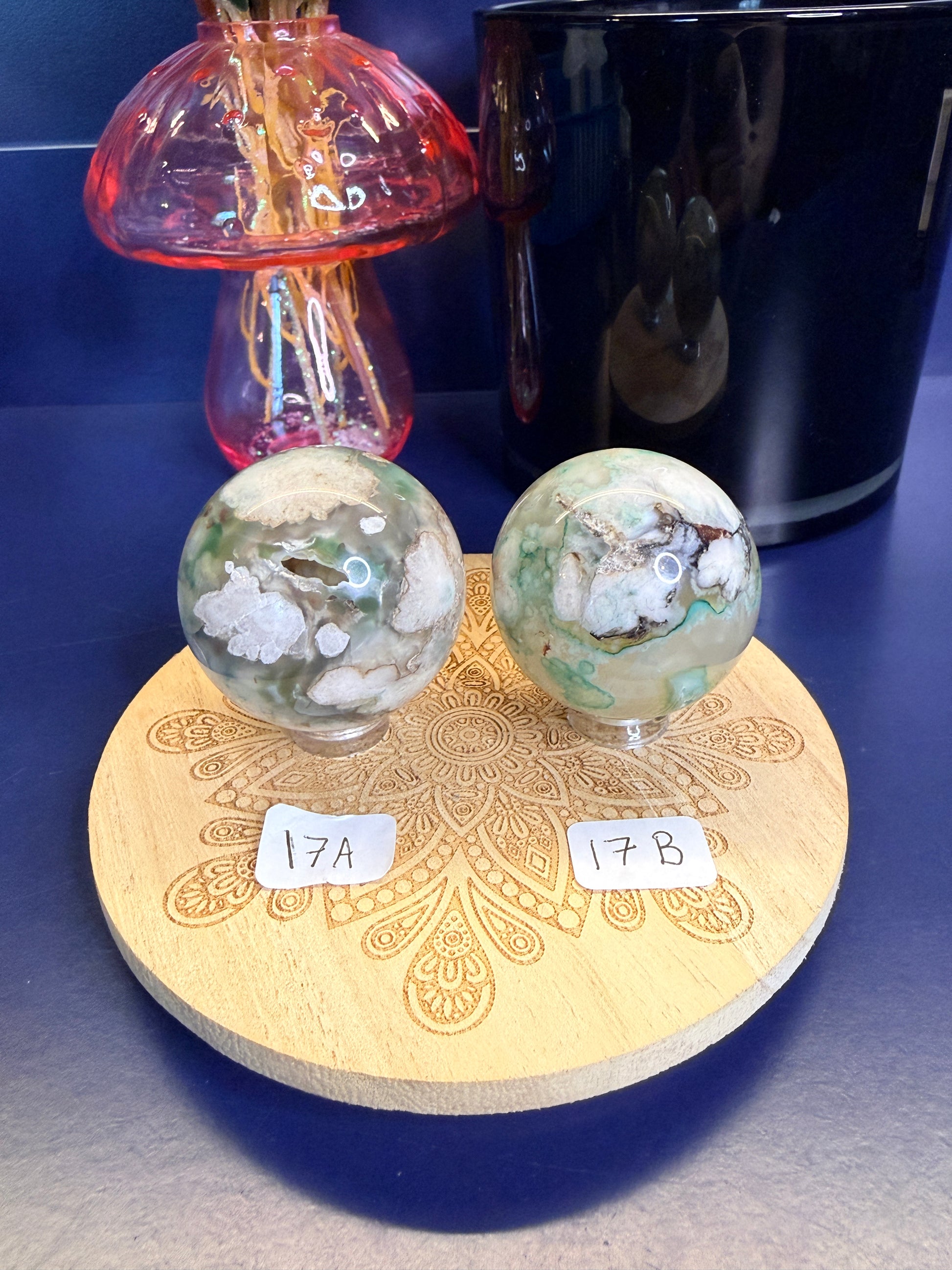 Two white and green translucent flower agate spheres on a wooden coaster in front of a black jar and pink mushroom