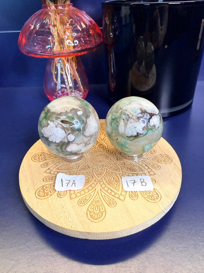 Two white and green translucent flower agate spheres on a wooden coaster in front of a black jar and pink mushroom
