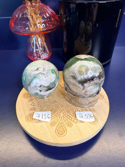 Two white and green translucent flower agate spheres on a wooden coaster in front of a black jar and pink mushroom