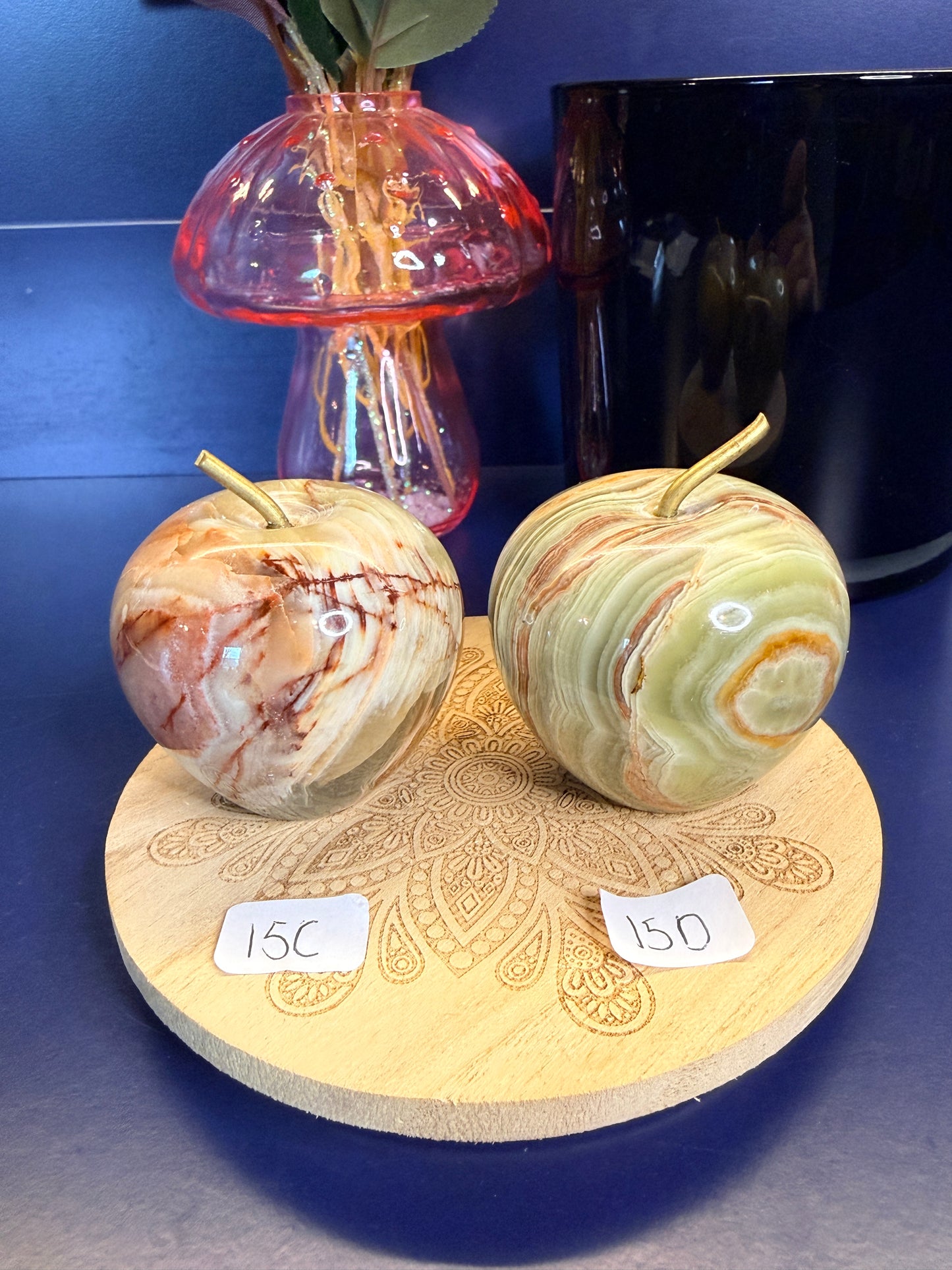 Two brown, red, and green onyx apples on a decorative wooden coaster in front of a mushroom vase and black jar