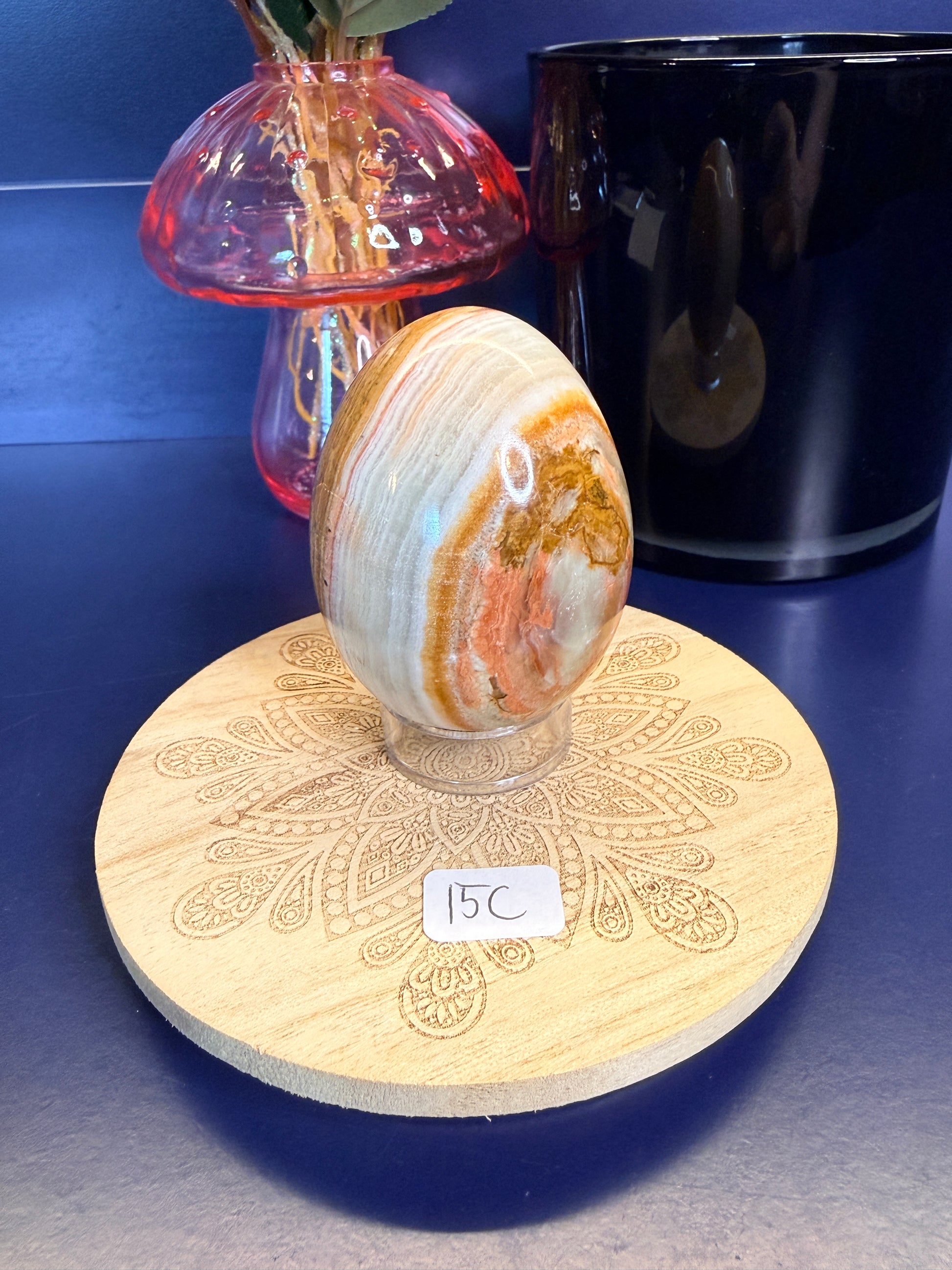 A brown, red, and green onyx egg on a decorative wooden coaster in front of a mushroom vase and black jar