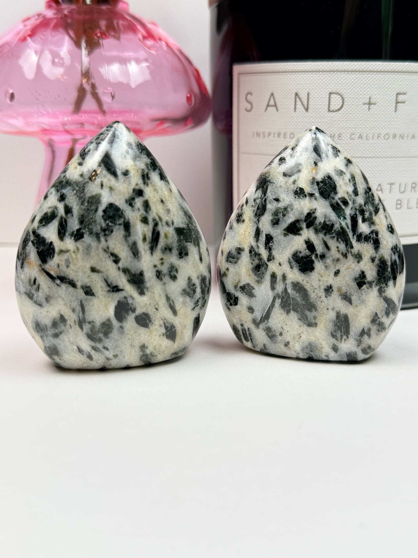 Two green tourmaline free forms in front of a black candle jar and pink vase on a white background