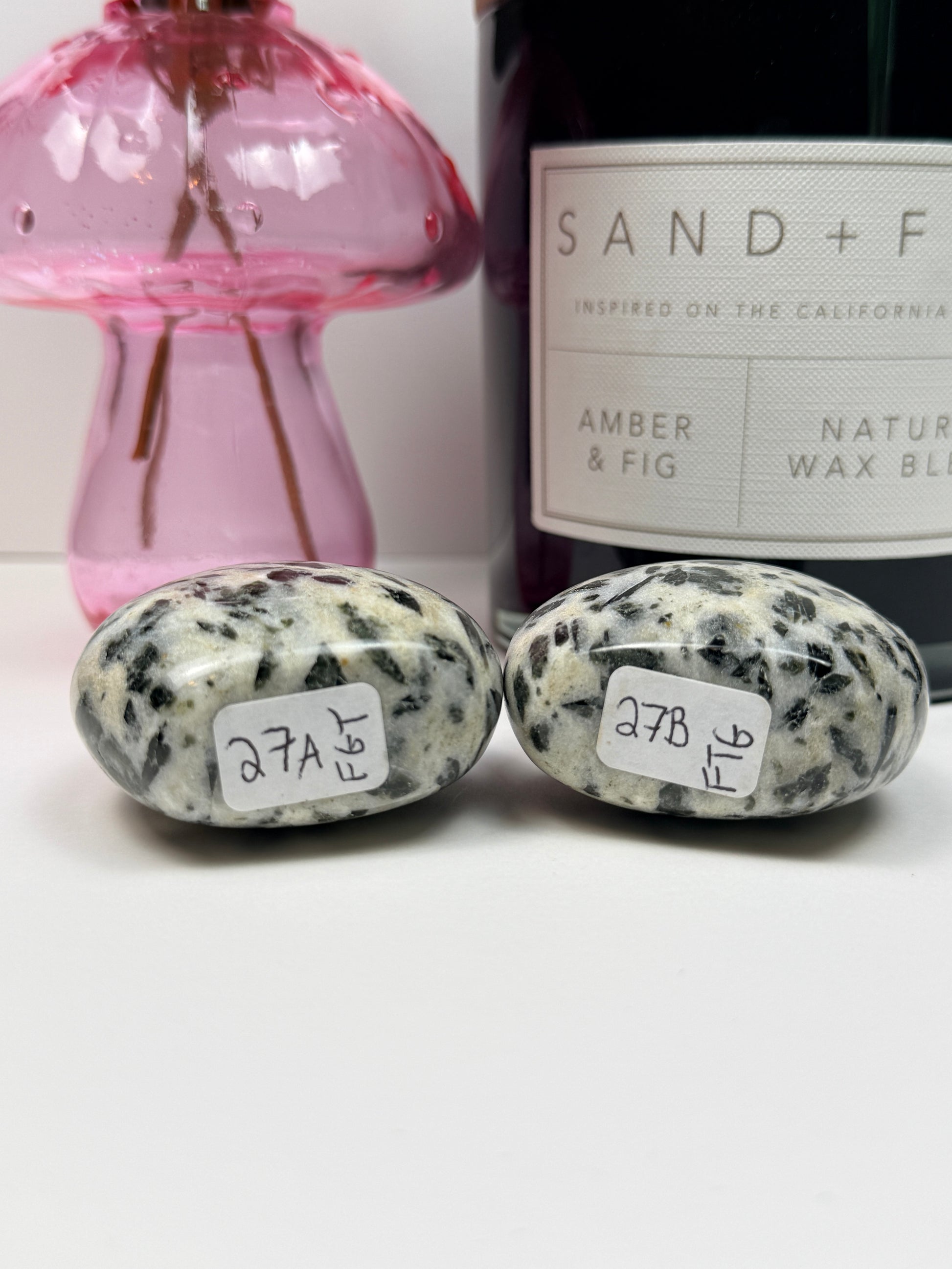 The bottom of two green tourmaline free forms with labels in front of a black candle jar and pink vase on a white background