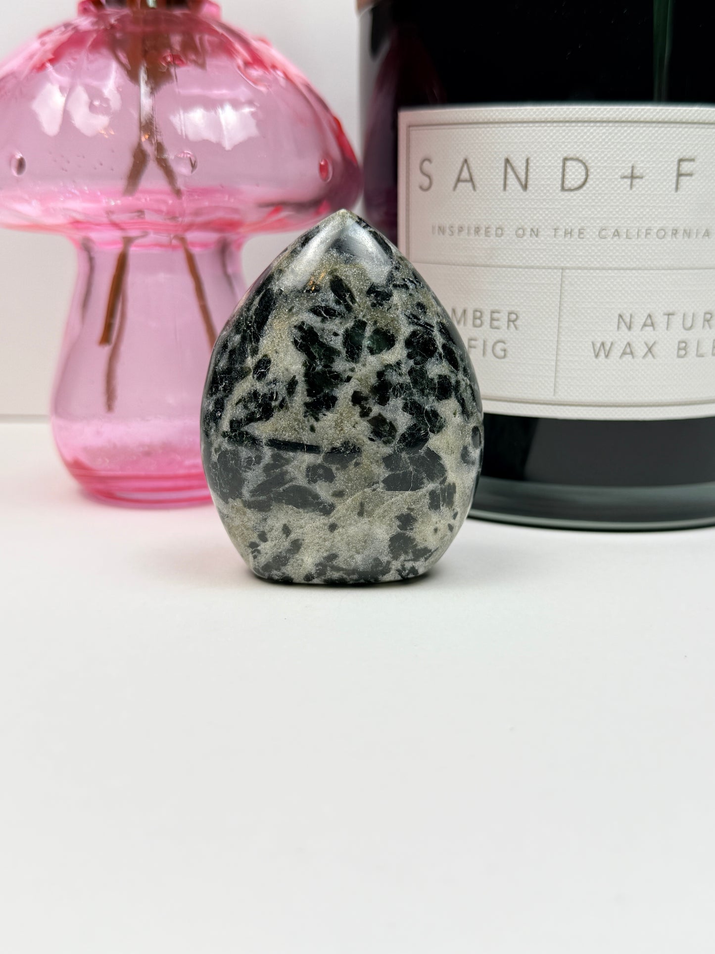 A polished green tourmaline free form in front of a black candle jar and pink vase on a white background