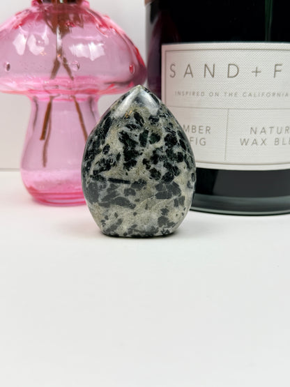 A polished green tourmaline free form in front of a black candle jar and pink vase on a white background