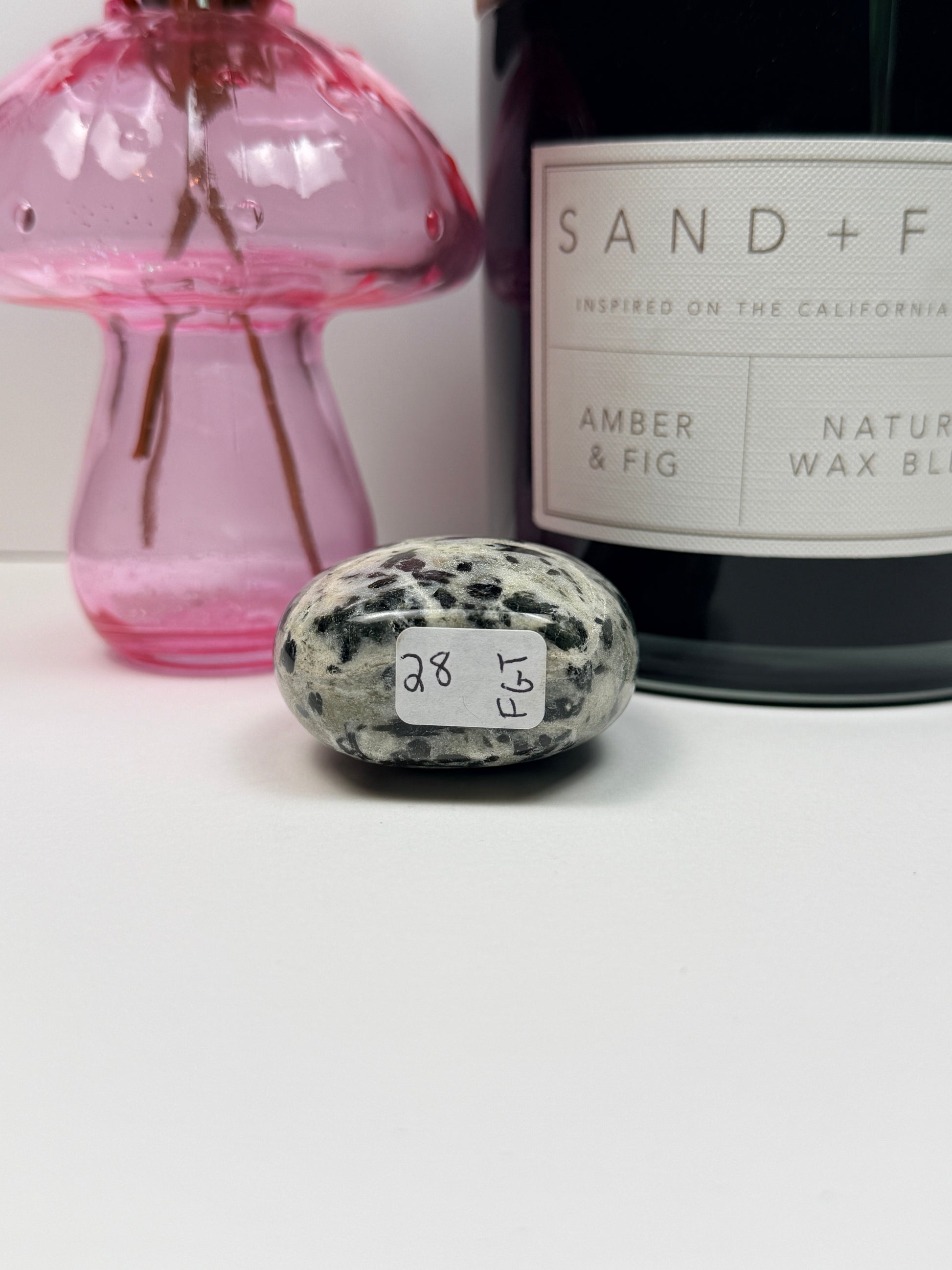 The bottom of a green tourmaline freeform with a '28' label in front of a black candle jar and pink vase on a white background