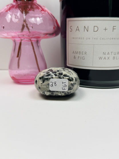 The bottom of a green tourmaline freeform with a '28' label in front of a black candle jar and pink vase on a white background