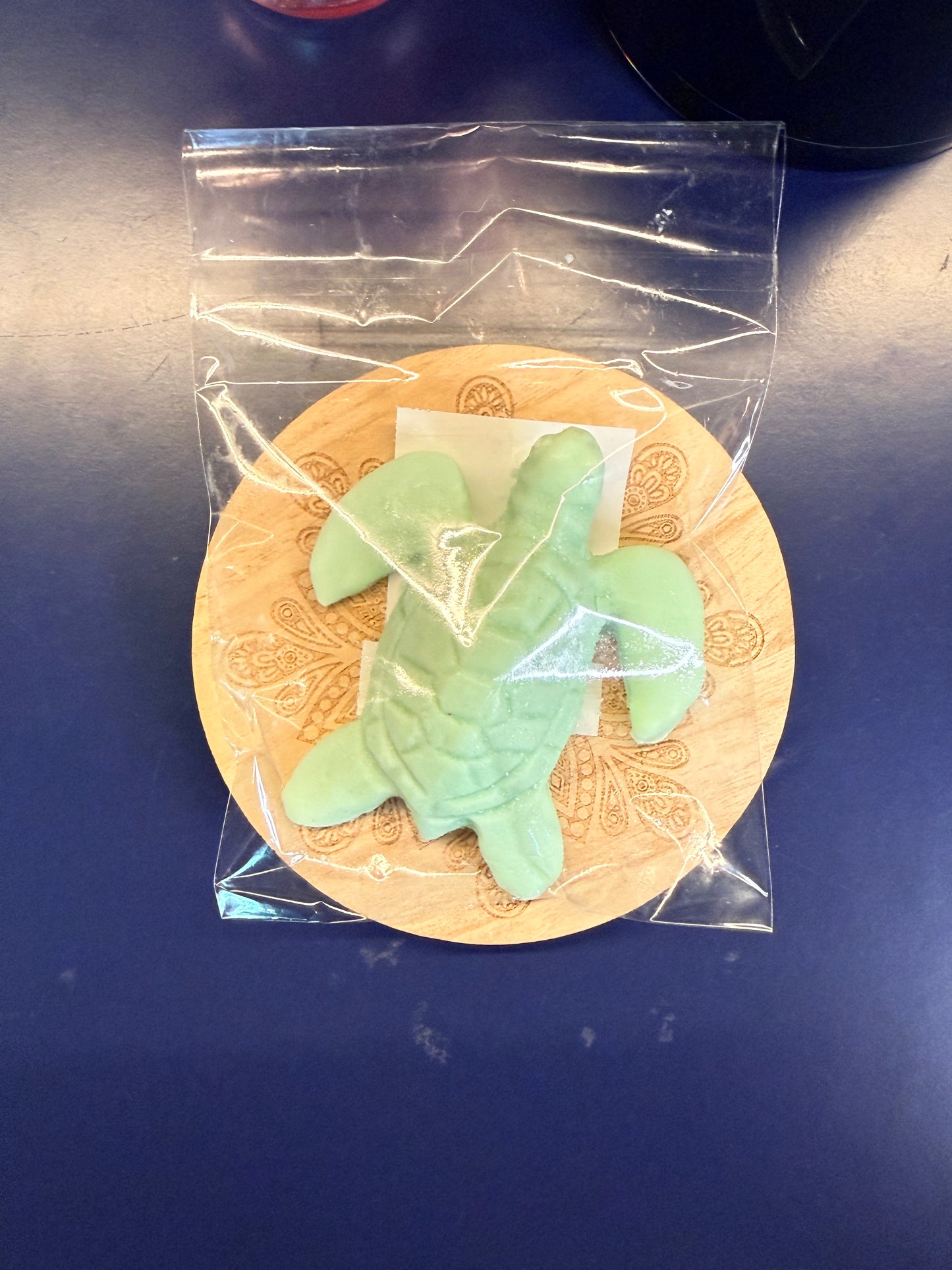 Green turtle soap bar on a wooden coaster, wrapped in clear plastic on a dark surface.