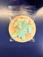 Green turtle soap bar on a wooden coaster, wrapped in clear plastic on a dark surface.