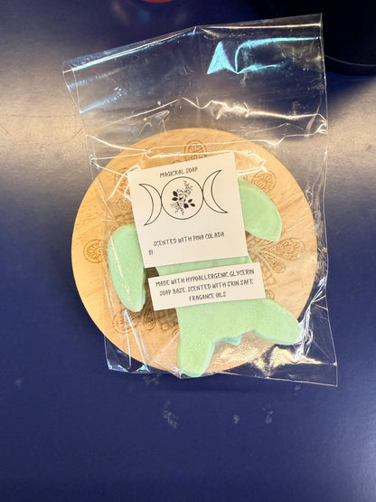 Green turtle soap bar with two labels on a wooden coaster, wrapped in clear plastic on a dark surface.