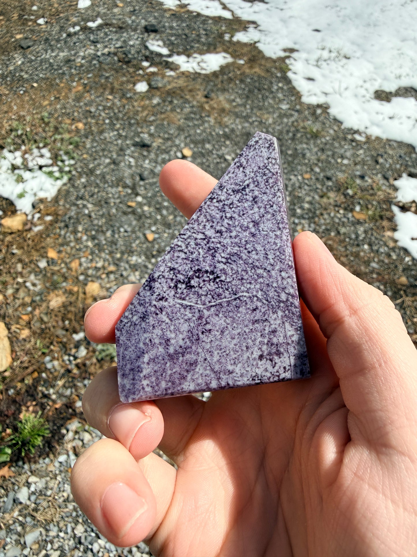 Hand holding a dark purple speckled kammererite freeform in front of gravel