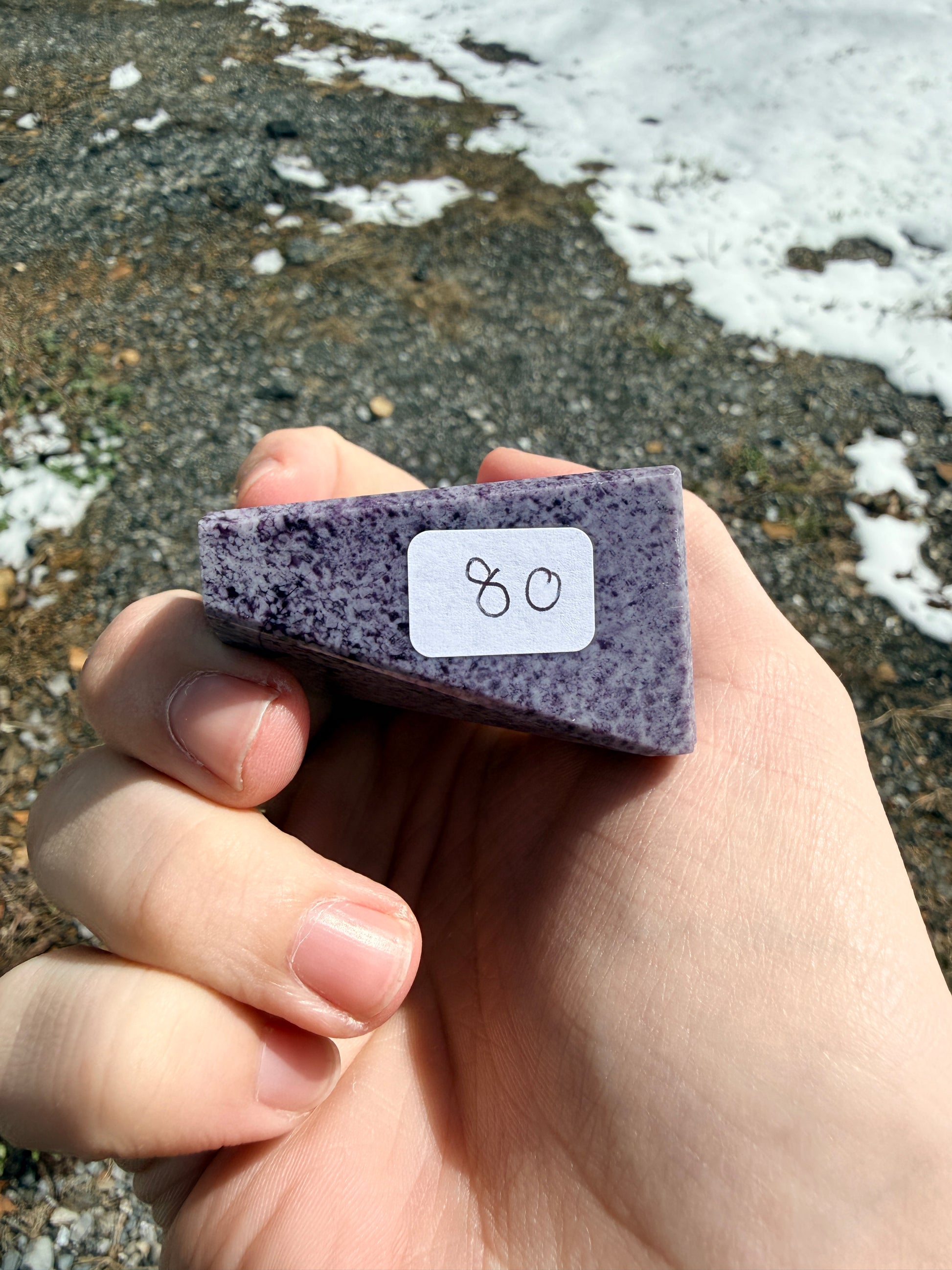 Hand holding a dark purple speckled kammererite freeform with an "80" label in front of gravel