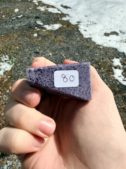Hand holding a dark purple speckled kammererite freeform with an "80" label in front of gravel