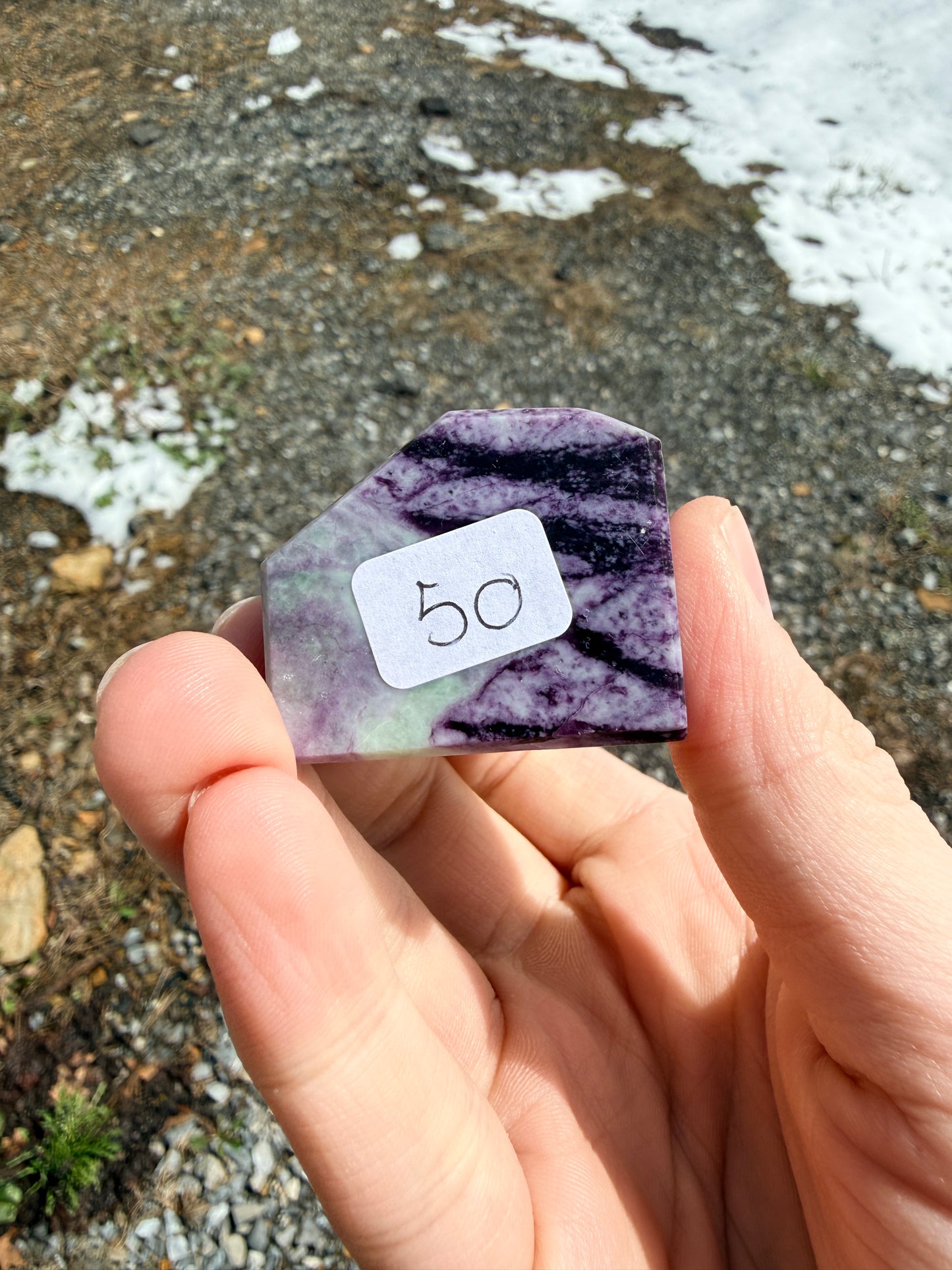 Hand holding a dark purple kammererite freeform with a "50" label in front of gravel