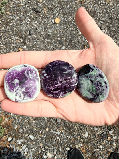 Hand holding three dark purple and light green kammererite palmstones with labels in front of gravel