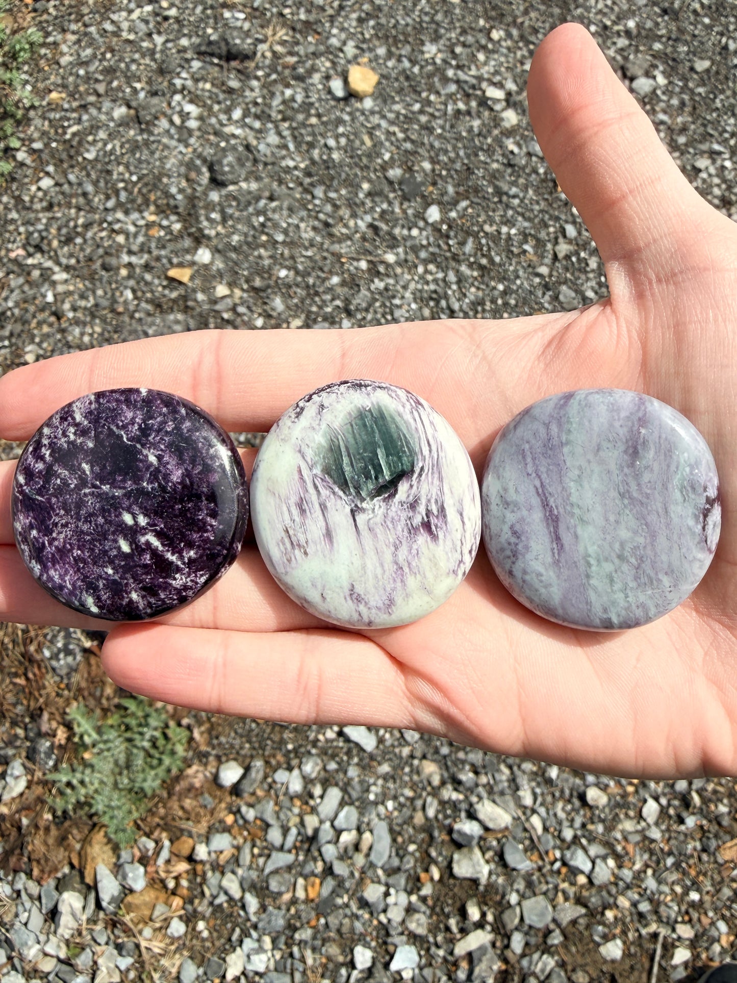 Hand holding three dark purple and light green kammererite palmstones in front of gravel