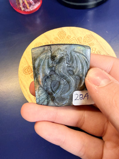 Hand holding a labradorite dragon slab with a yellow flash in front of a wooden coaster against a blue background