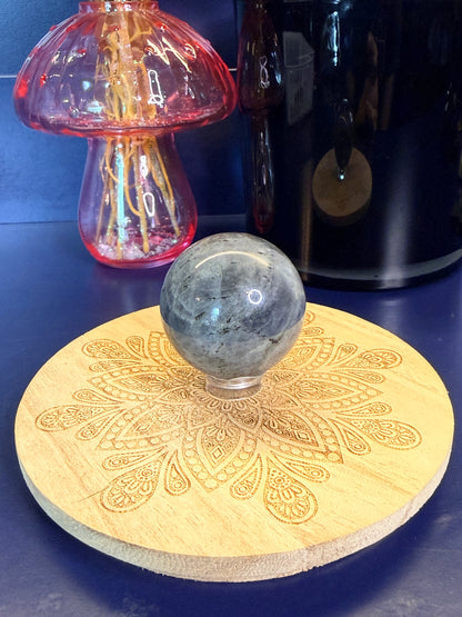 Grey patterned labradorite sphere sitting on a decorative wooden coaster in front of a pink mushroom and black jar