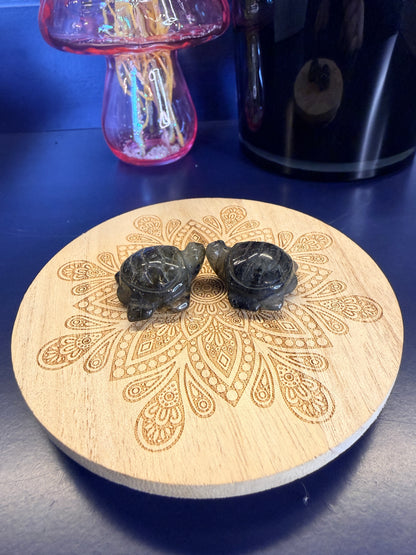 Two grey and black crystal turtles on a decorative wooden coaster in front of a vase and black jar