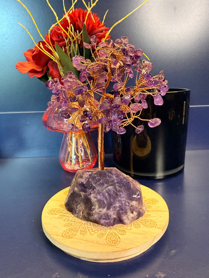 A purple crystal chip tree made of copper wire in a large amethyst crystal on a wooden coaster in front of a pink mushroom and black jar