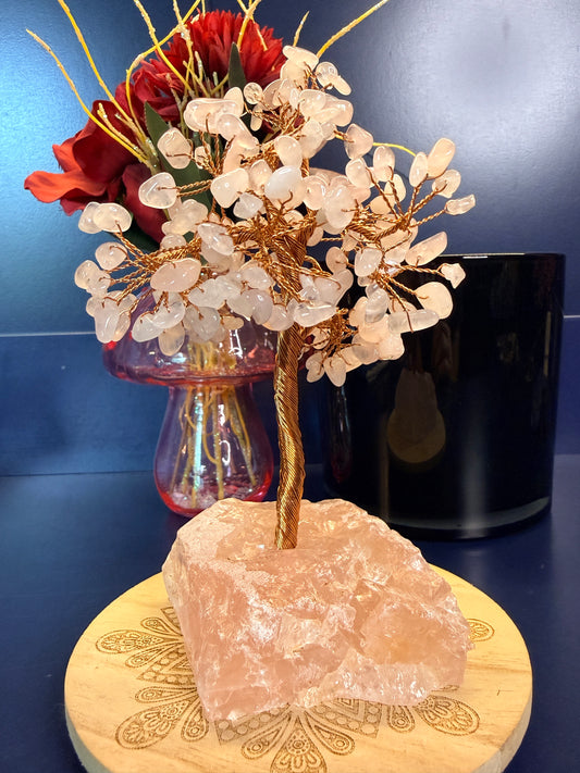 A pink crystal chip tree made of copper wire in a large rose quartz crystal on a wooden coaster in front of a pink mushroom and black jar