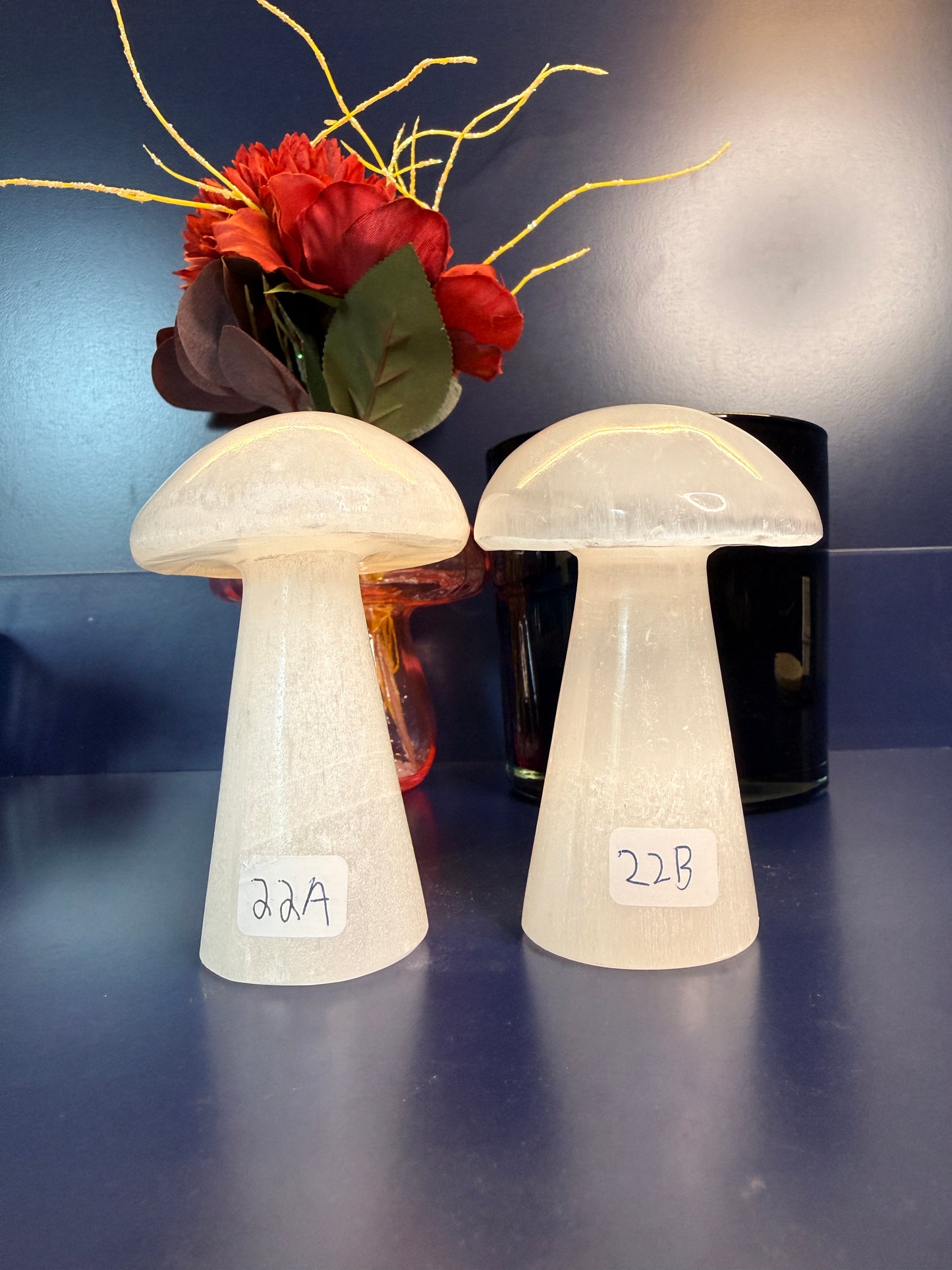 Two white mushroom-shaped selenite in front of a red flower and black jar on a blue background