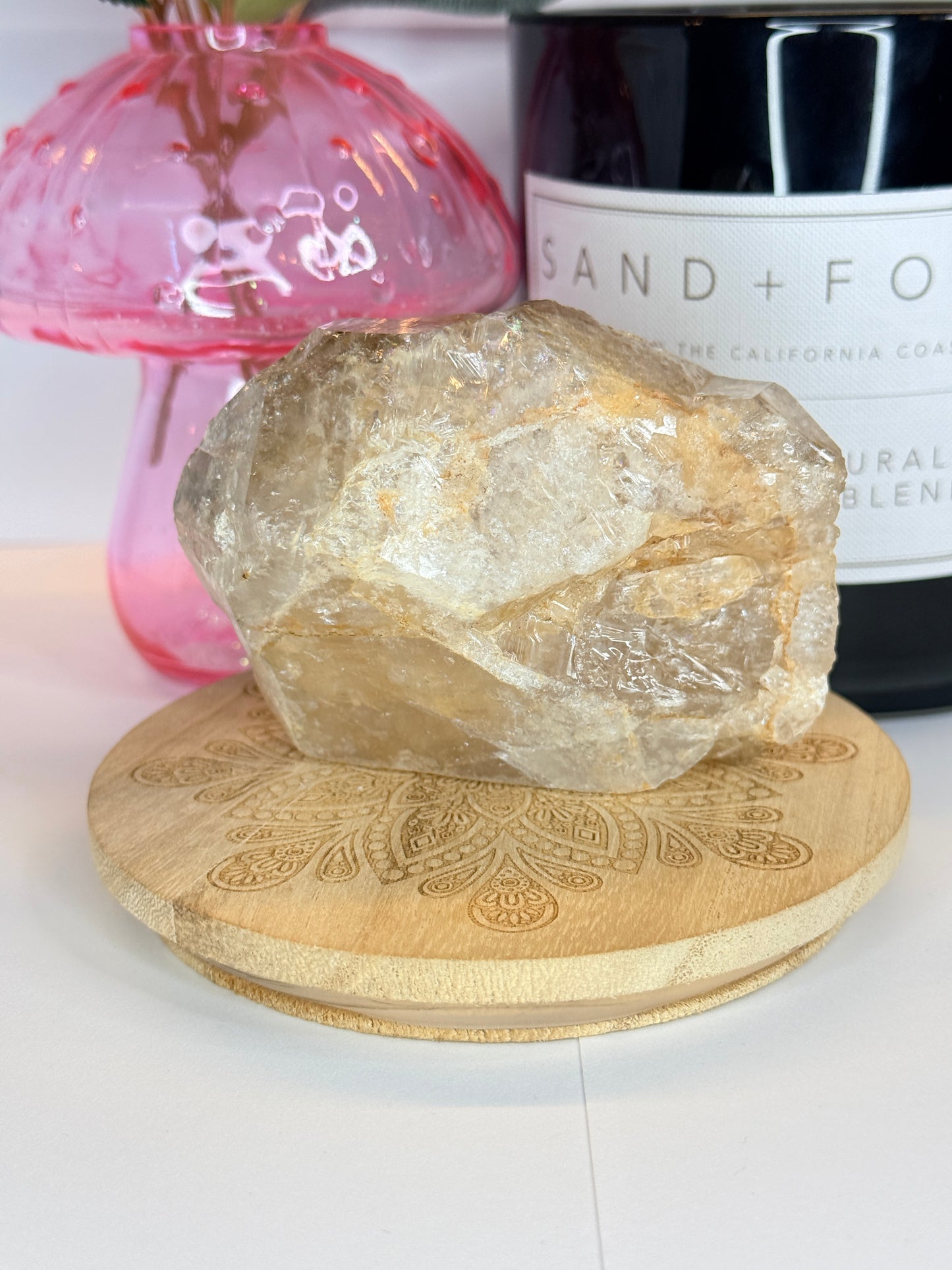 Large raw smokey quartz specimen resting on a decorative wooden coaster in front of a black candle jar and pink vase on a white background