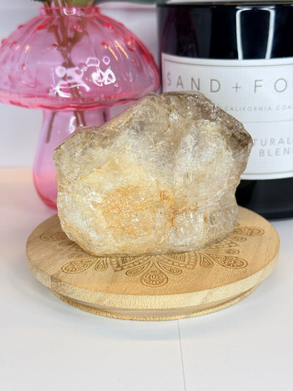 A large smokey quartz raw specimen resting on a decorative wooden coaster in front of a black candle jar and pink vase on a white background