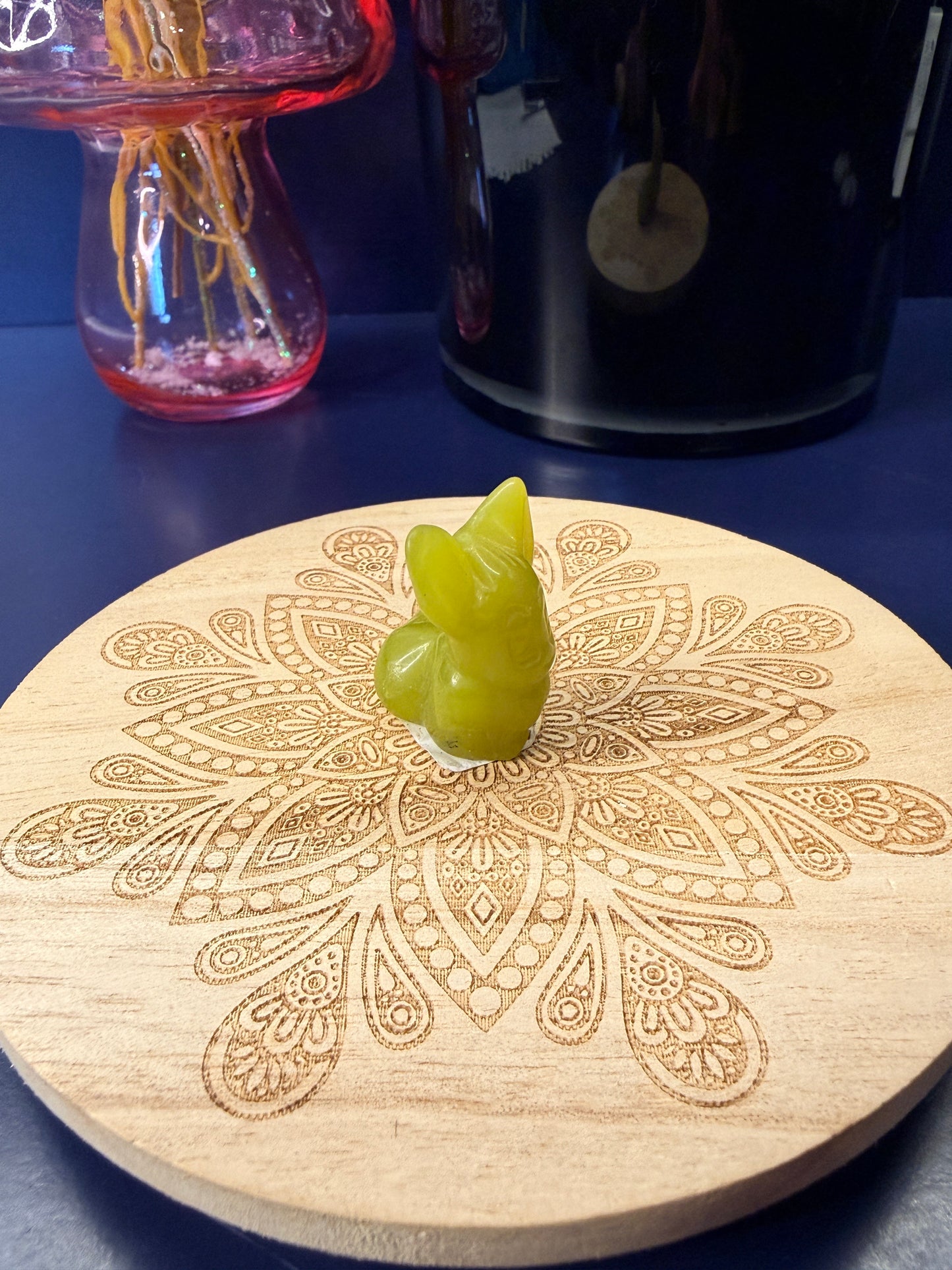Lemon Calcite eevee crystal shape sitting on a decorative wooden coaster in front of a glass mushroom and black jar
