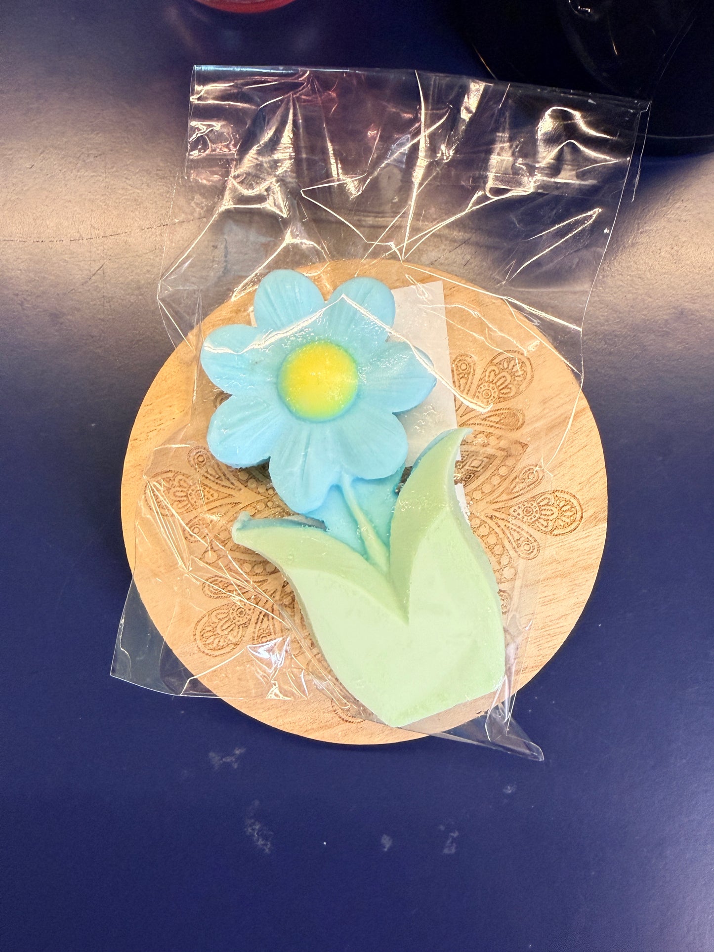 Blue and green flower soap bar on a wooden coaster, wrapped in clear plastic on a dark surface.