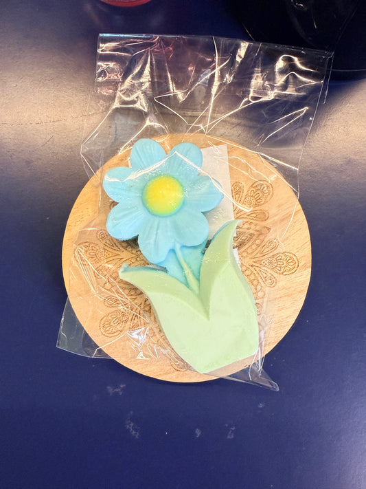 Blue and green flower soap bar on a wooden coaster, wrapped in clear plastic on a dark surface.