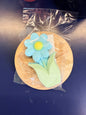 Blue and green flower soap bar on a wooden coaster, wrapped in clear plastic on a dark surface.