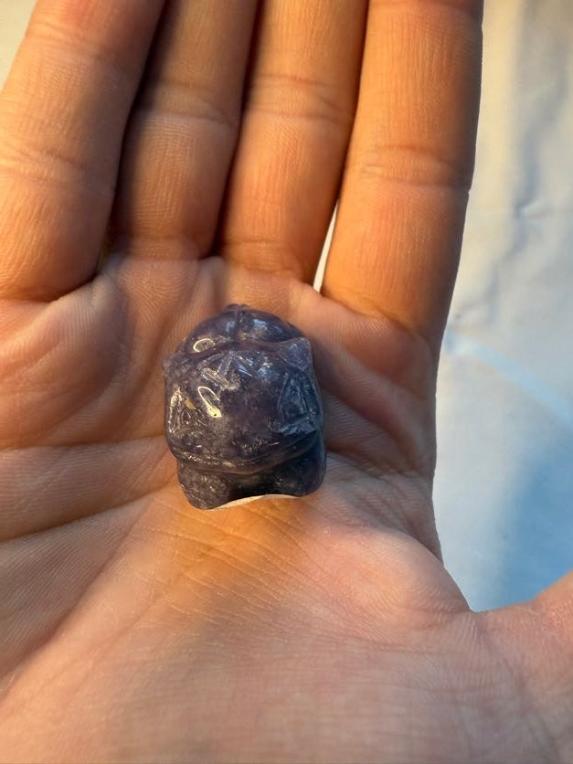 Hand holding a purple lepidolite crystal carving of a bulbasaur-inspired creature against a white background