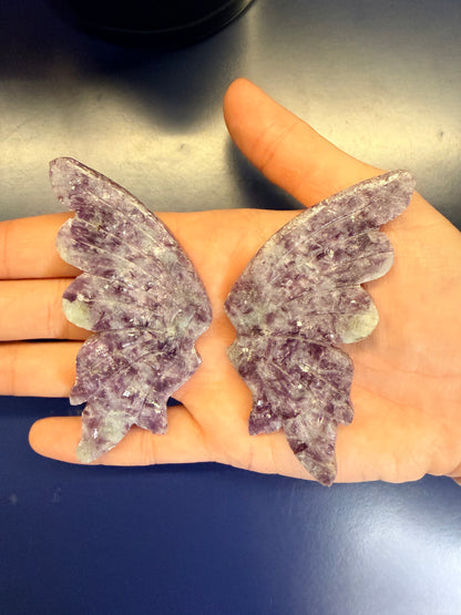 Hand holding two large purple lepidolite butterfly wings against a blue background