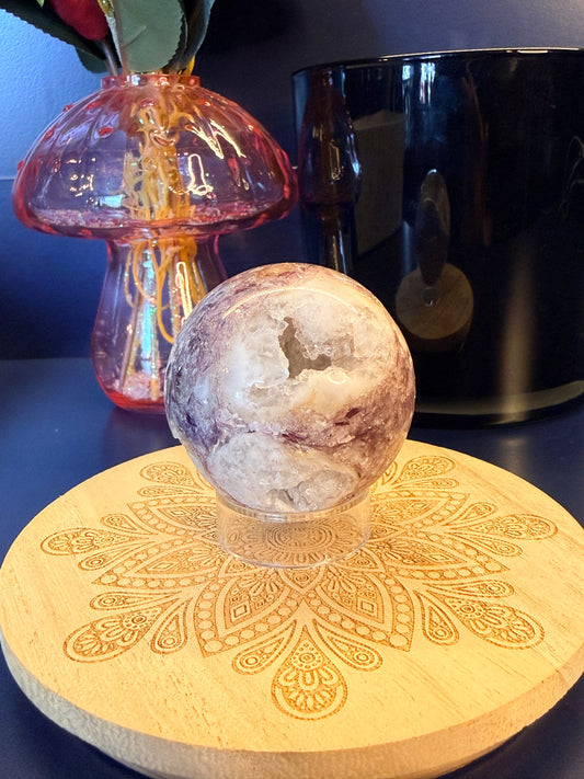 A white and purple crystal sphere sitting on a decorative wooden coaster in front of a glass mushroom and black jar